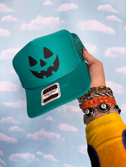 A hand wearing colorful bracelets holds a teal trucker hat with a black jack-o'-lantern face graphic. The hat has a white and red 'Gildan' brand patch on the brim, set against a light blue sky with white clouds backdrop.