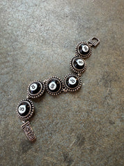 An 8-ball themed bracelet featuring five circular black and white 8-billiard motif links surrounded by ornate bronze-toned bezels, with a matching rectangular charm and clasp, displayed on a textured gray stone surface.