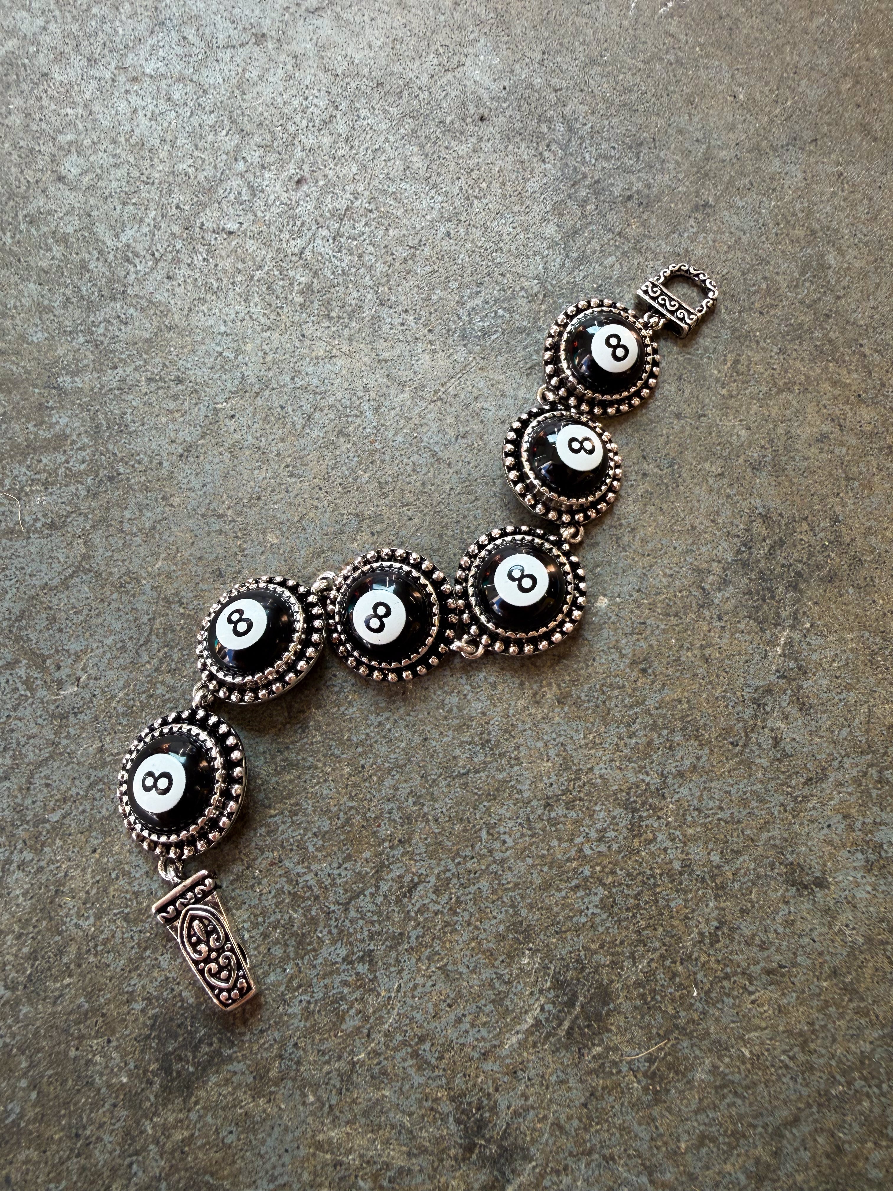 An 8-ball themed bracelet featuring five circular black and white 8-billiard motif links surrounded by ornate bronze-toned bezels, with a matching rectangular charm and clasp, displayed on a textured gray stone surface.