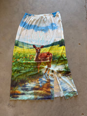 A long, wide-leg skirt with a colorful landscape print featuring a deer in a field and water, laid flat on a concrete floor.