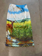 A long, wide skirt or sarong laid flat on a concrete floor, featuring a vibrant, painterly landscape print with a deer, green foliage, blue sky, and yellow fields