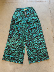 A pair of wide-leg cropped pants with a vibrant turquoise and black leopard print pattern, laid flat on a concrete floor. The brand name 'cocoon' is visible on the hanging tag