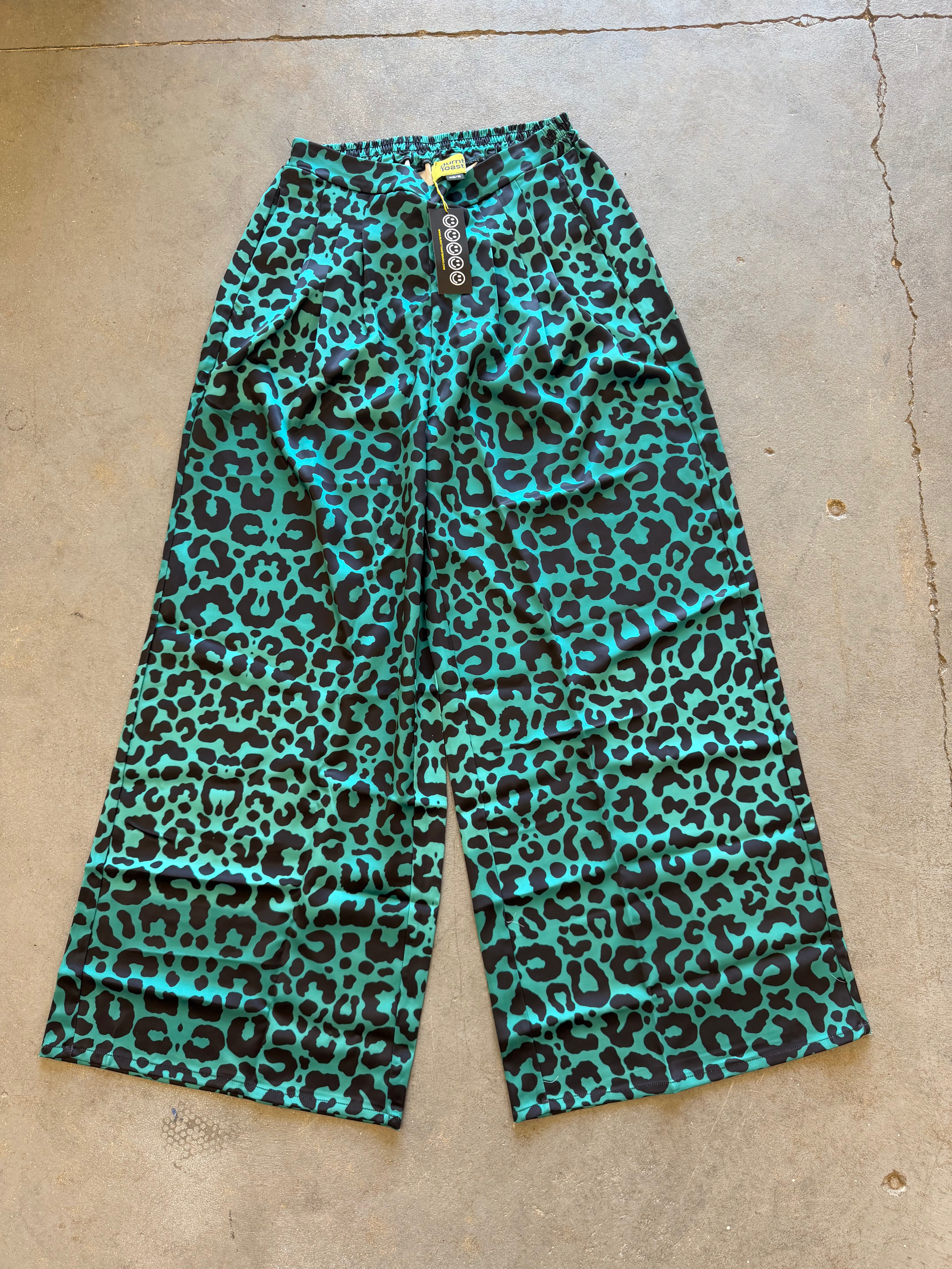 A pair of wide-leg cropped pants with a vibrant turquoise and black leopard print pattern, laid flat on a concrete floor. The brand name 'cocoon' is visible on the hanging tag