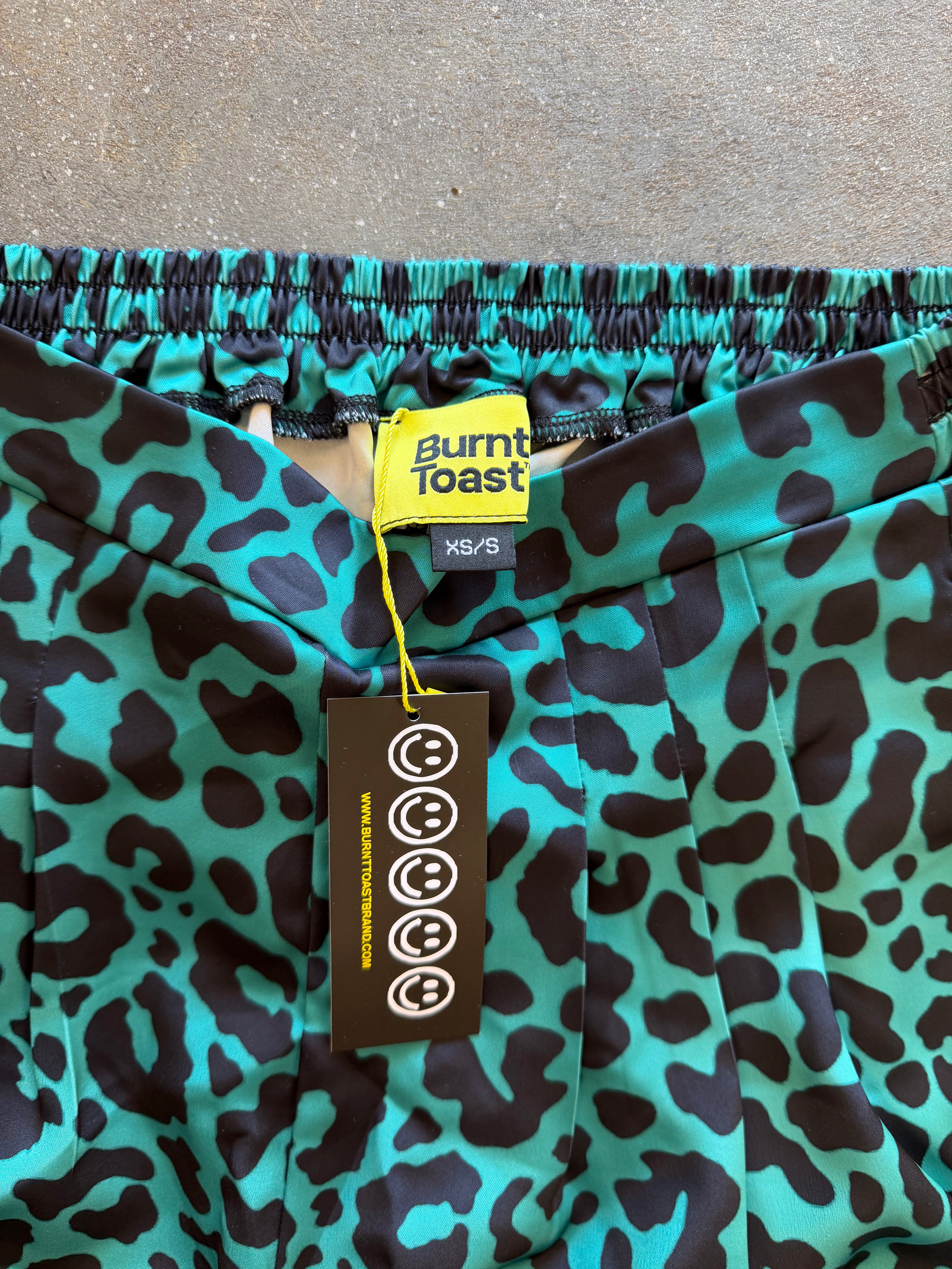 Close-up of a pair of turquoise and black leopard-print pants with an elastic waistband, featuring a yellow XS/S Burnt Toast brand tag