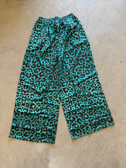 A pair of wide-leg cropped pants with a vibrant turquoise and black leopard print, laid flat on a beige concrete floor