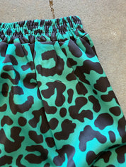 Close-up of a pair of leopard-print shorts or pants in bright turquoise green with dark brown spots, featuring an elastic waistband, laid flat on a concrete surface