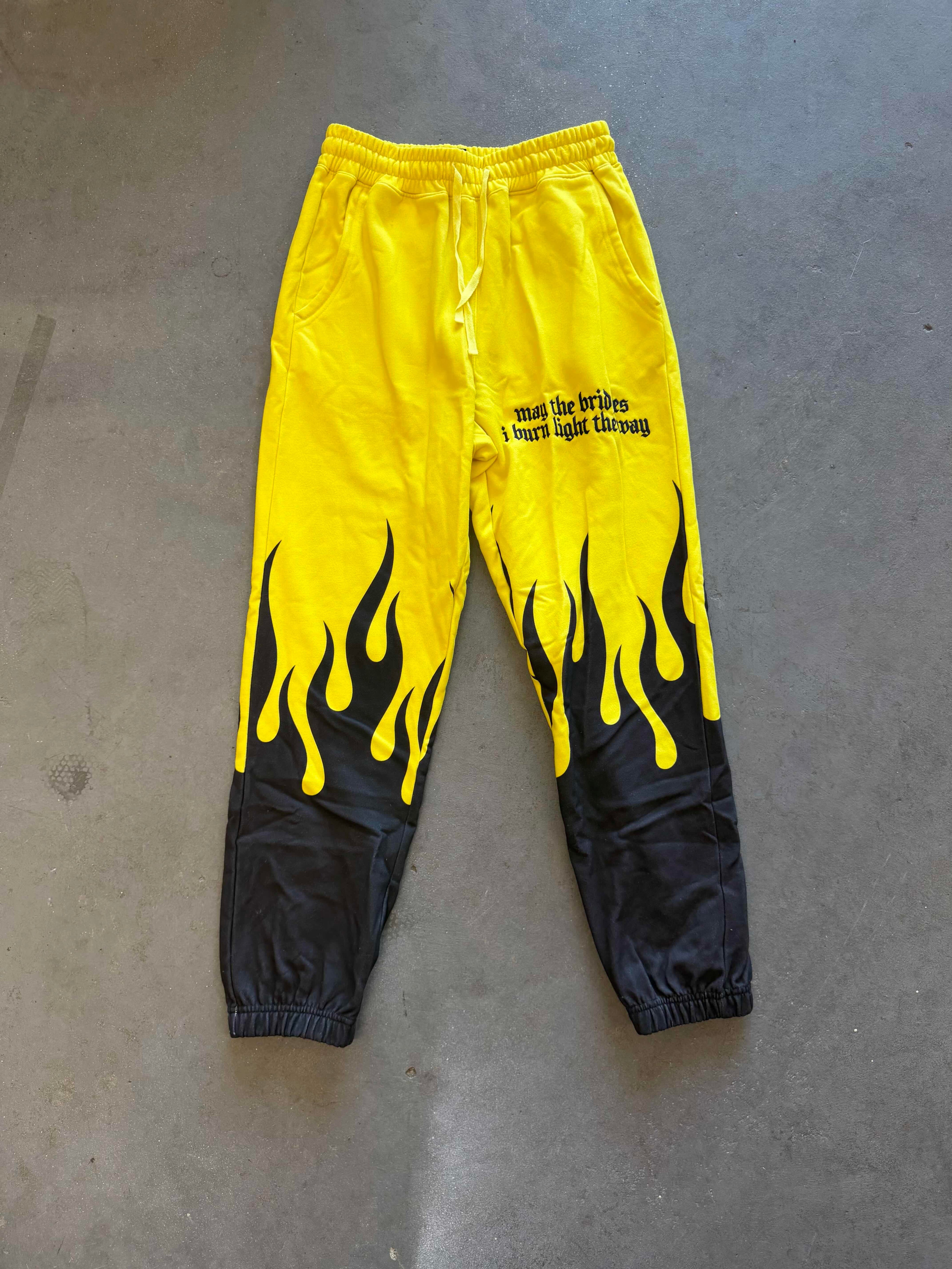 Bright yellow and black flame-print sweatpants laid flat on a gray concrete floor, featuring black elastic cuffs and a small black text graphic on the right leg