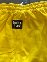 Close-up of bright yellow sweatpants with an elastic waistband and a black rectangular patch reading 'burnt toast' in white gothic lettering
