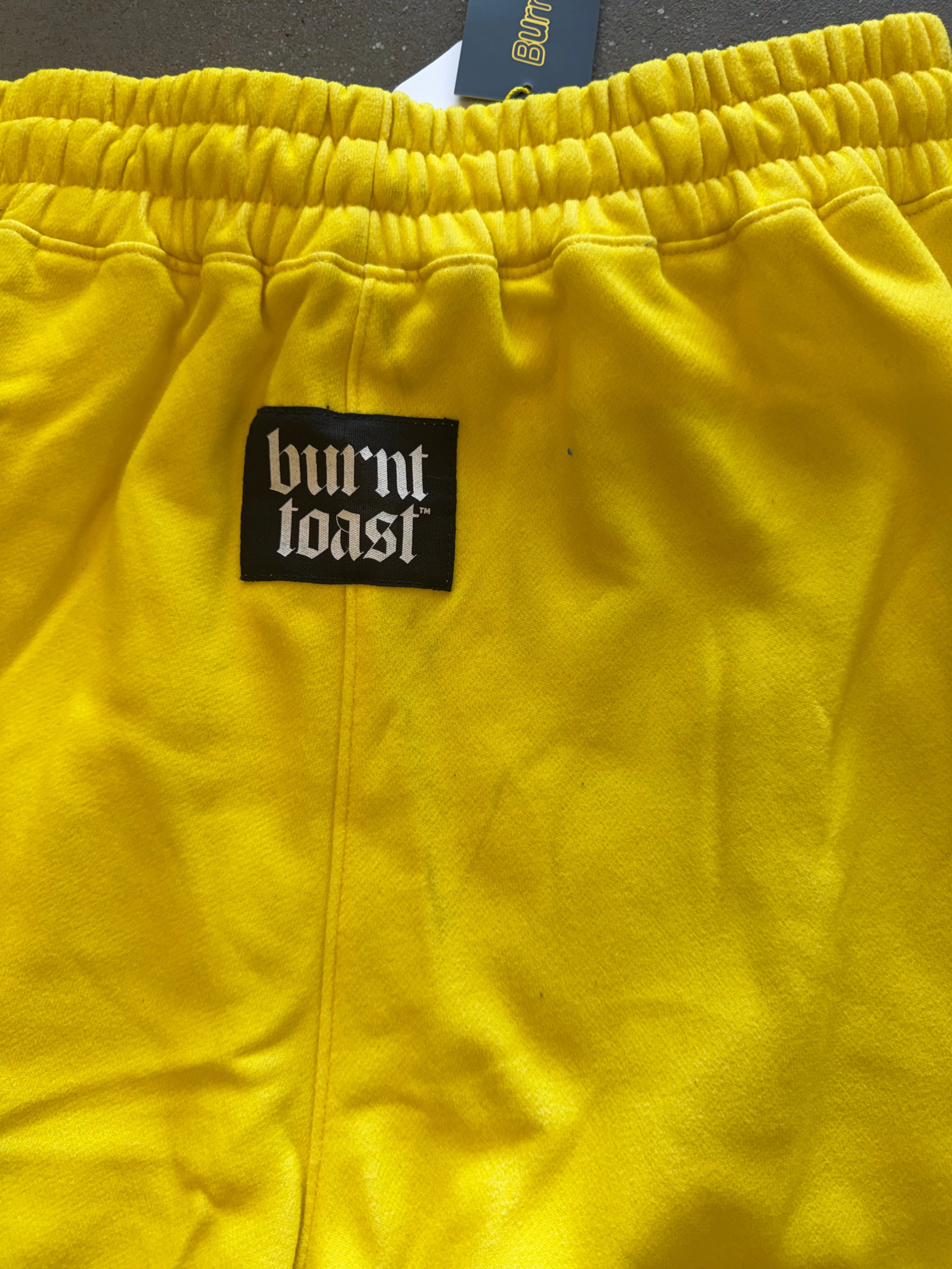 Close-up of bright yellow sweatpants with an elastic waistband and a black rectangular patch reading 'burnt toast' in white gothic lettering
