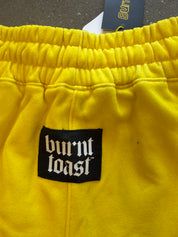 Close-up of bright yellow sweatpants with an elastic waistband, a black Burnt Toast brand patch on the front, and a hanging Burnt Toast tag