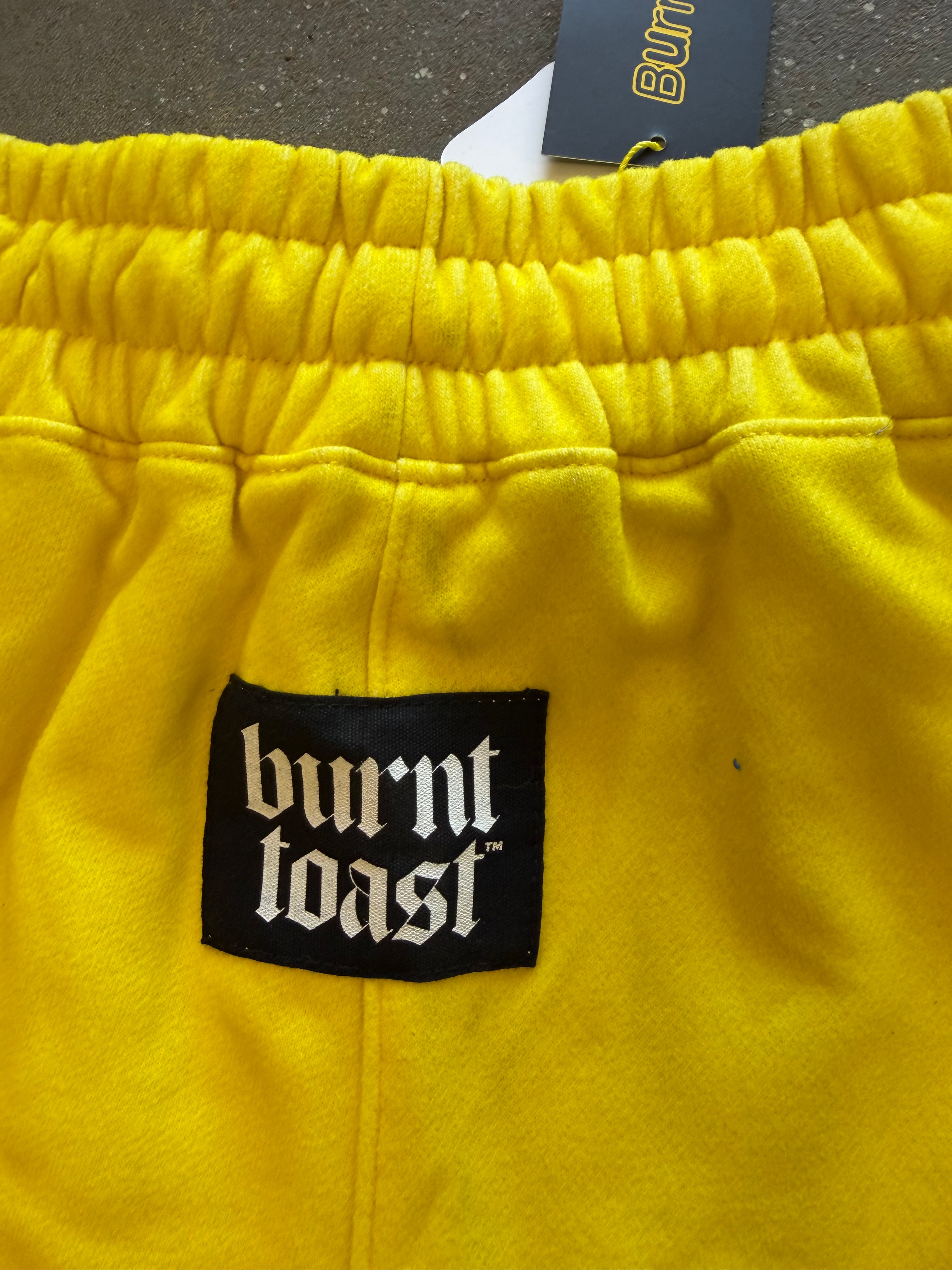 Close-up of bright yellow sweatpants with an elastic waistband, a black Burnt Toast brand patch on the front, and a hanging Burnt Toast tag