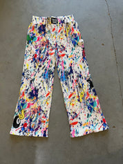 A pair of wide-leg pleated pants featuring a vibrant multicolor paint splatter and splodge pattern on a white base, laid flat on a gray concrete floor