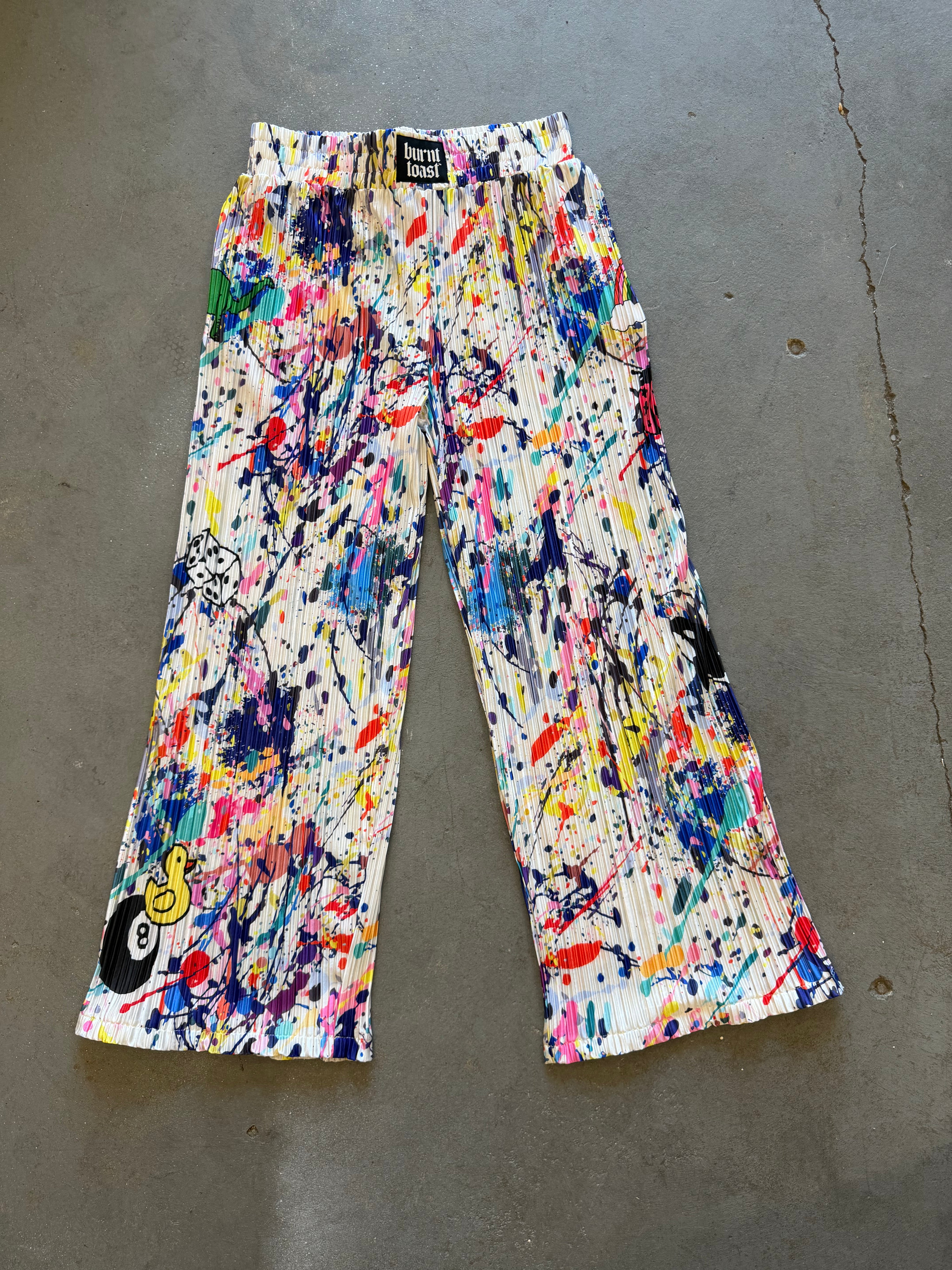 A pair of wide-leg pleated pants featuring a vibrant multicolor paint splatter and splodge pattern on a white base, laid flat on a gray concrete floor