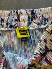 Close-up of a Burnt Toast garment with a colorful paint-splatter pattern, featuring a yellow brand label and elasticized ruffled trim