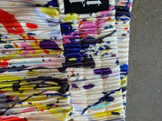 A close-up of a pleated fabric garment, likely a skirt or shorts, featuring a vibrant multicolor paint-splatter pattern on a white base. A black brand label with white arrows is visible at the top edge