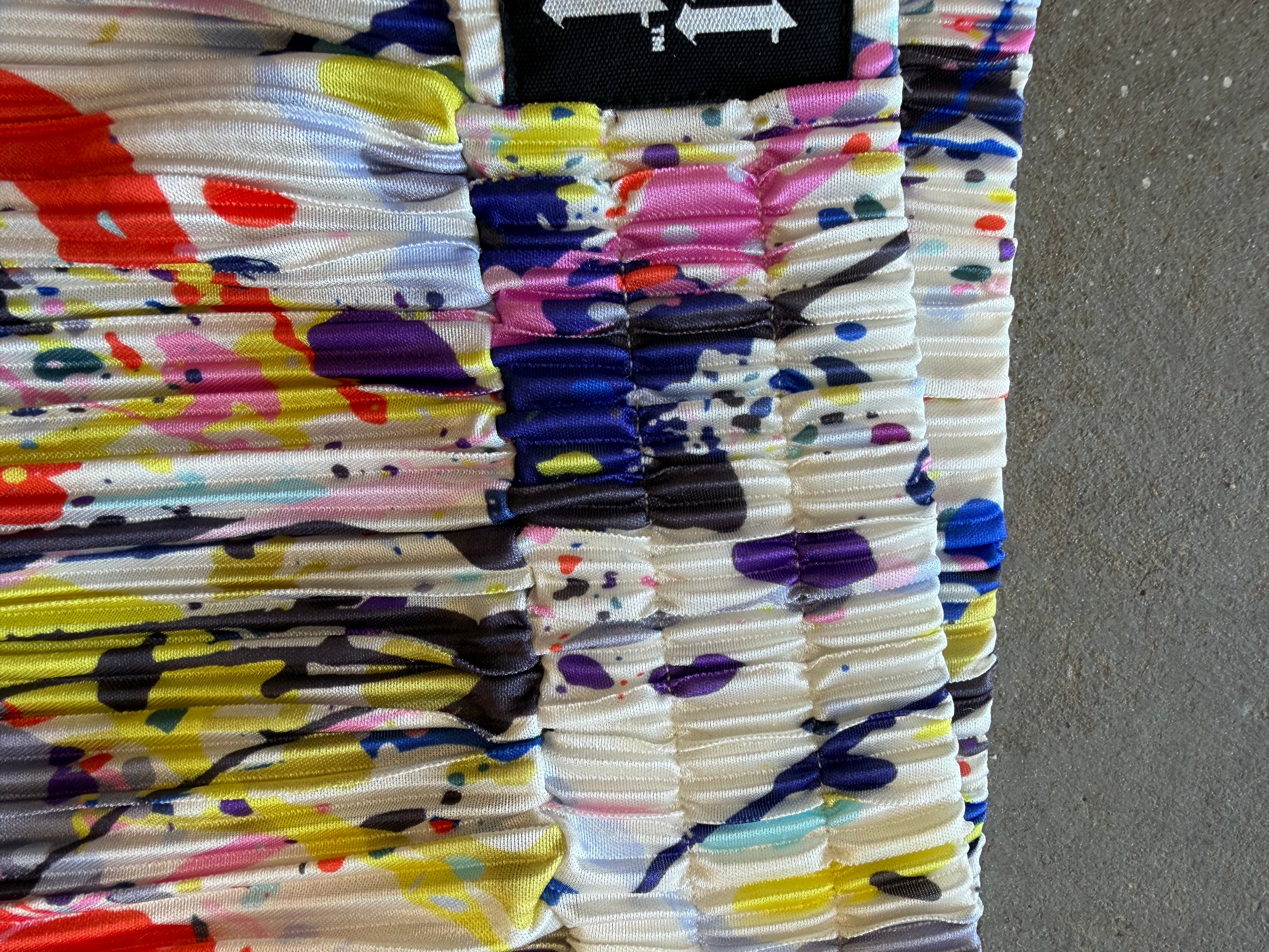 A close-up of a pleated fabric garment, likely a skirt or shorts, featuring a vibrant multicolor paint-splatter pattern on a white base. A black brand label with white arrows is visible at the top edge