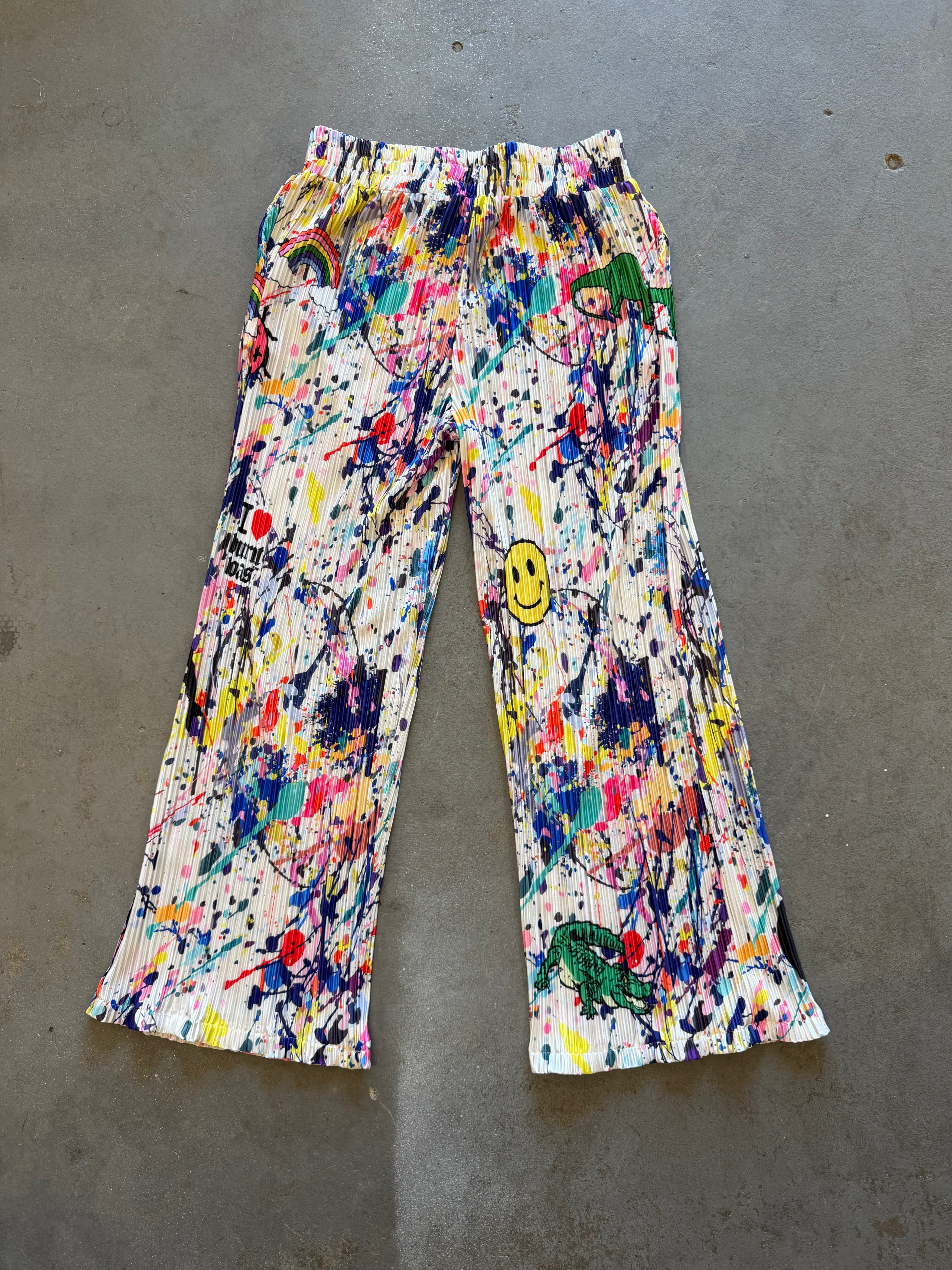 A pair of wide-leg cropped pants featuring a vibrant multicolor paint splatter and graffiti print on a white base, laid flat on a gray concrete floor