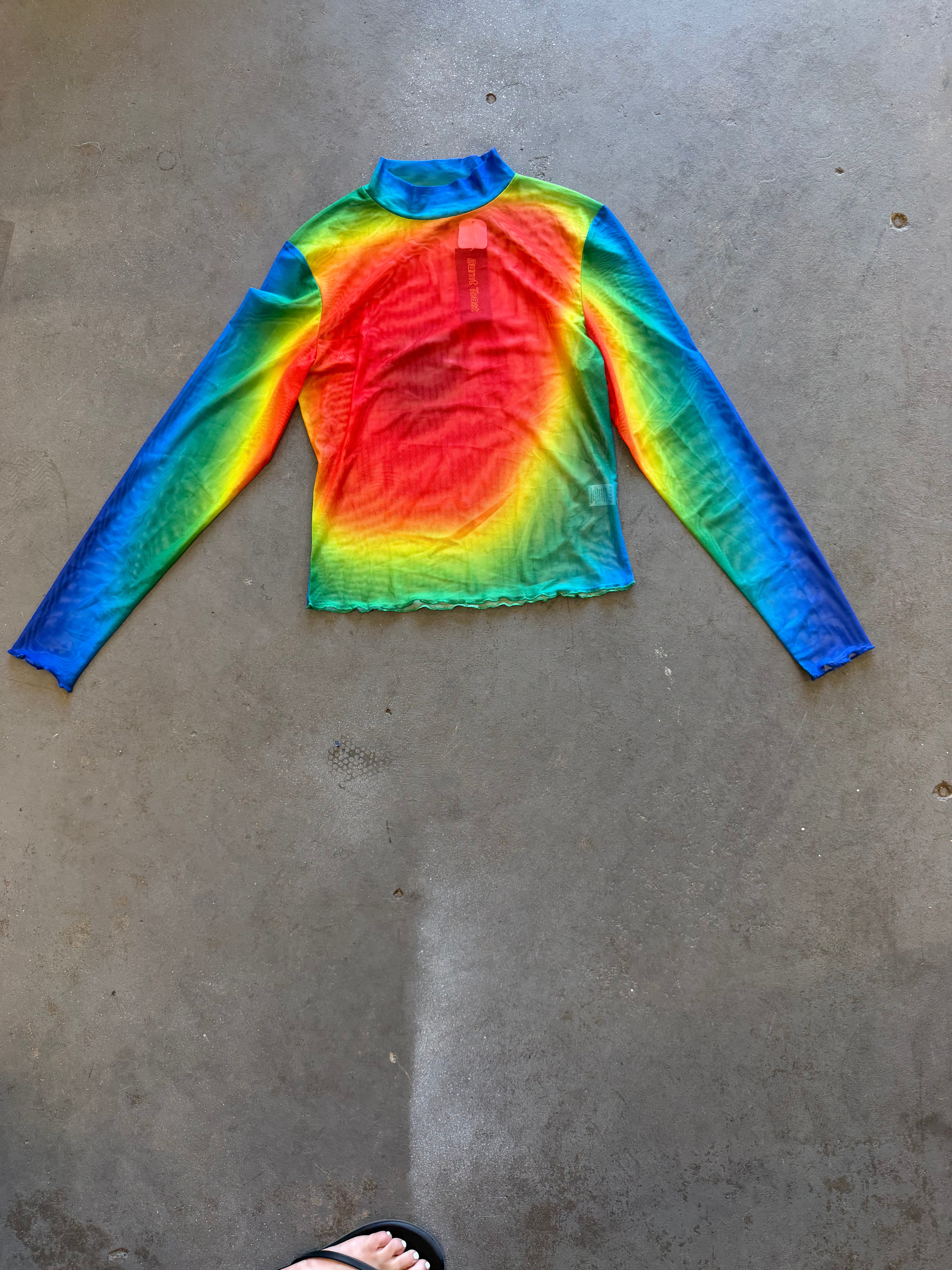 A long-sleeve mock neck shirt with a vibrant rainbow gradient pattern, laid flat on a gray concrete floor. A person's foot wearing a black sandal is visible at the bottom edge