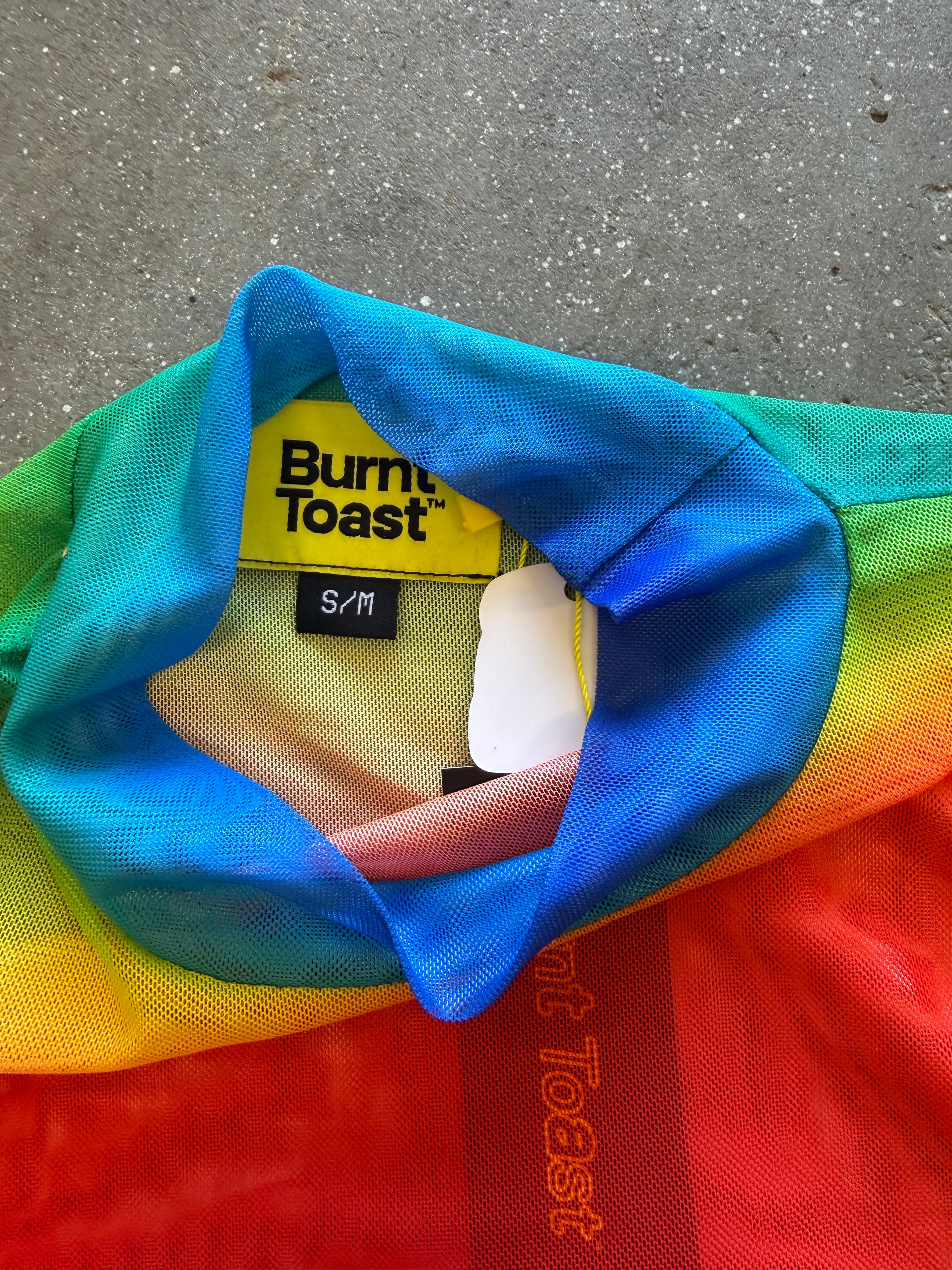 Close-up of a rainbow-striped mesh garment with a yellow Burne Toast brand label, laid on a gray concrete surface