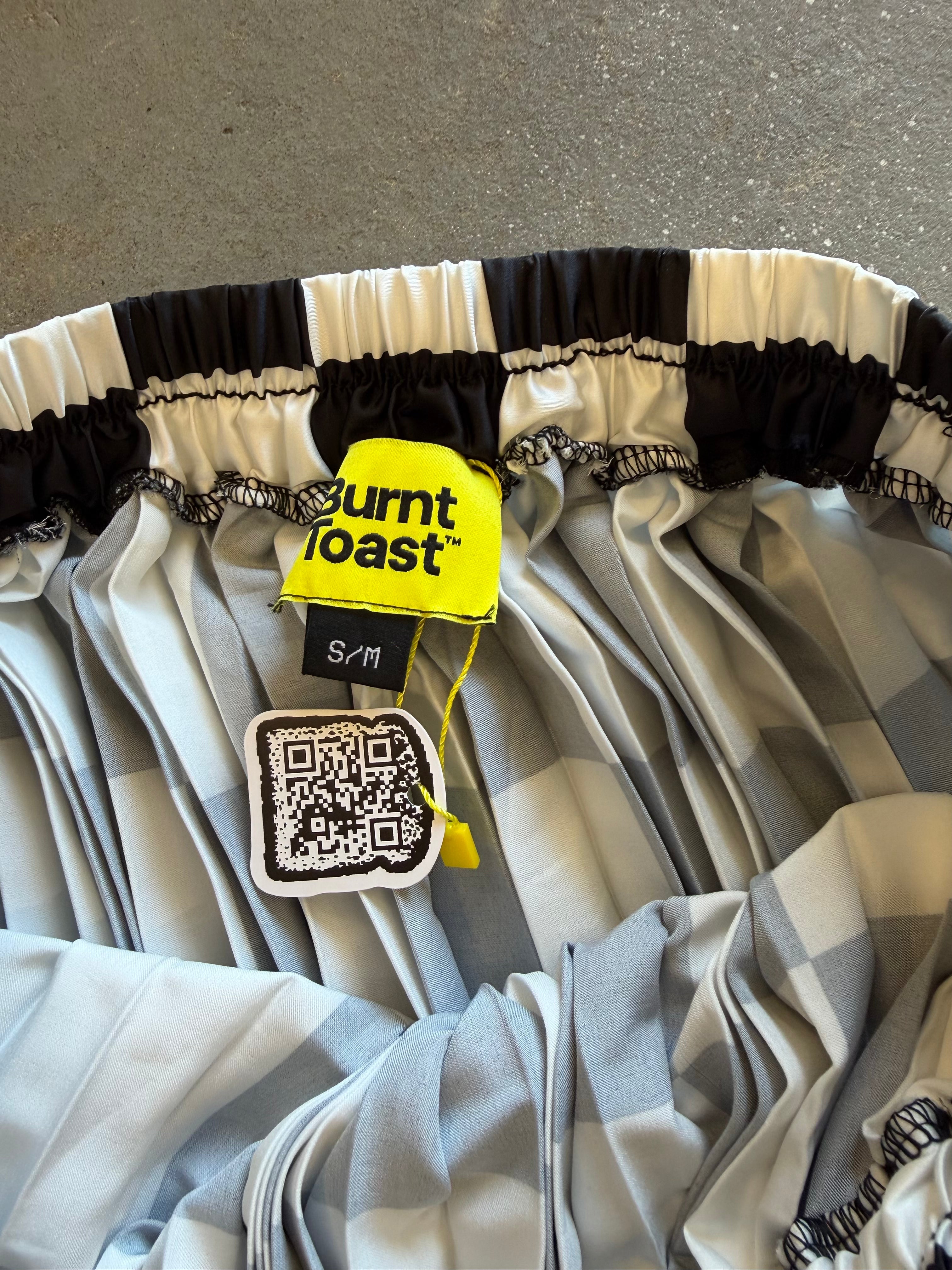 Close-up of a pleated skirt with a black and white striped ruffled waistband and a light gray and white plaid body, showing a yellow Burnt Toast brand tag and a QR code hangtag