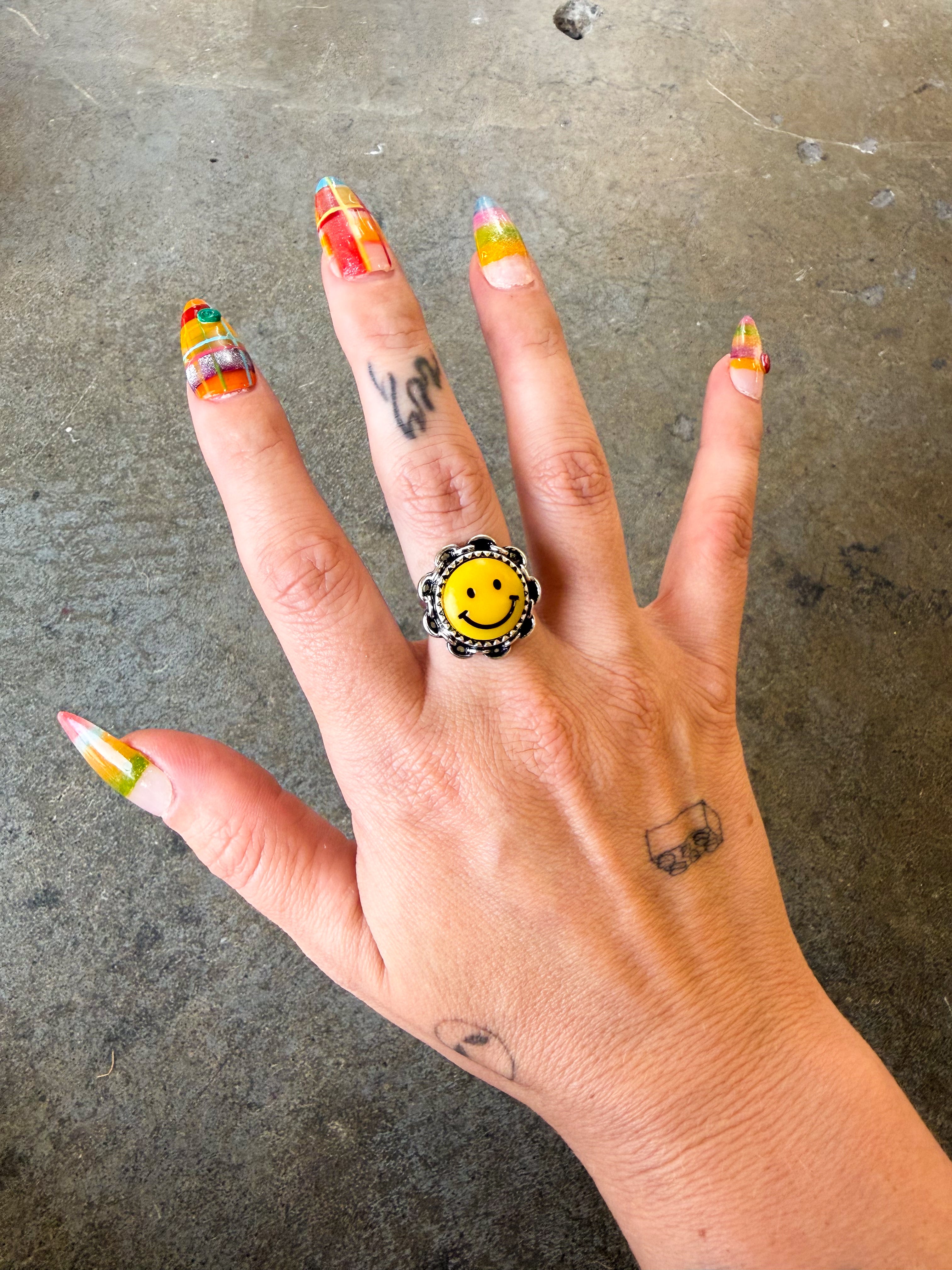A hand with colorful nail art and small tattoos wearing a chunky black ring with a yellow smiley face center, set against a concrete floor background.