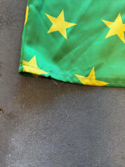 A close-up of a green fabric item, likely a flag or textile panel, featuring a repeating pattern of yellow five-pointed stars, laid flat on a dark gray concrete surface