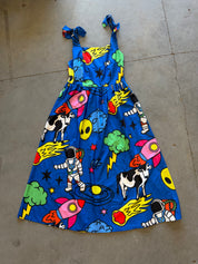 A sleeveless blue dress with a colorful retro space print featuring astronauts, cows, rockets, stars, and planets, laid flat on a concrete floor