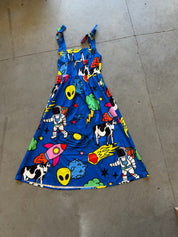 A blue sleeveless dress with a colorful space-themed print featuring astronauts, aliens, rockets, planets, and black-and-white cows, laid flat on a concrete floor