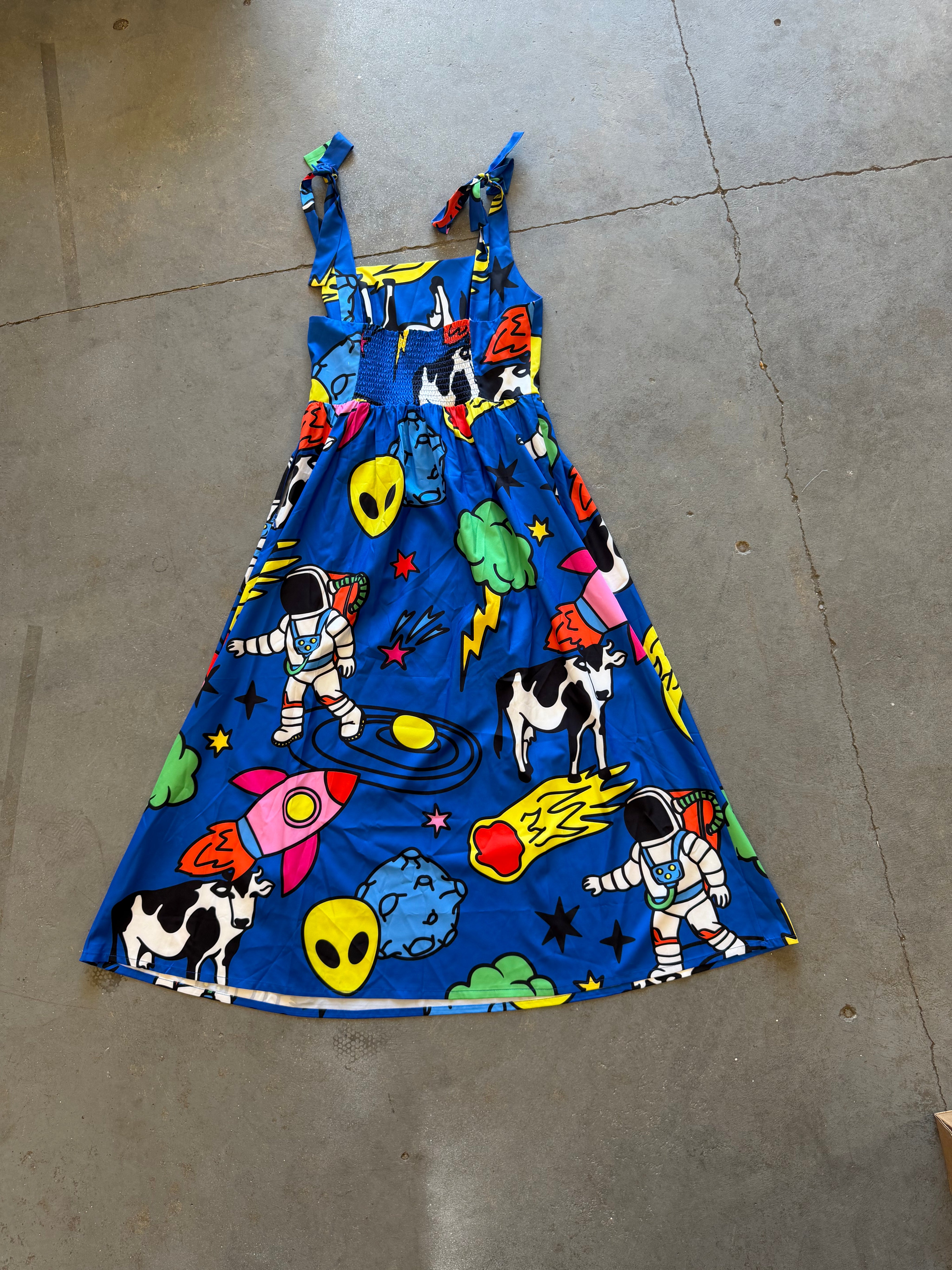 A blue sleeveless dress with a colorful space-themed print featuring astronauts, aliens, rockets, planets, and black-and-white cows, laid flat on a concrete floor