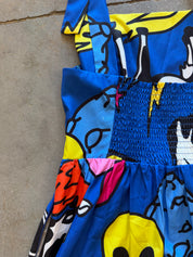 A close-up of a colorful printed garment laid flat on a concrete surface, featuring bold blue, yellow, black, pink, and orange abstract graphic patterns with a smocked waist section