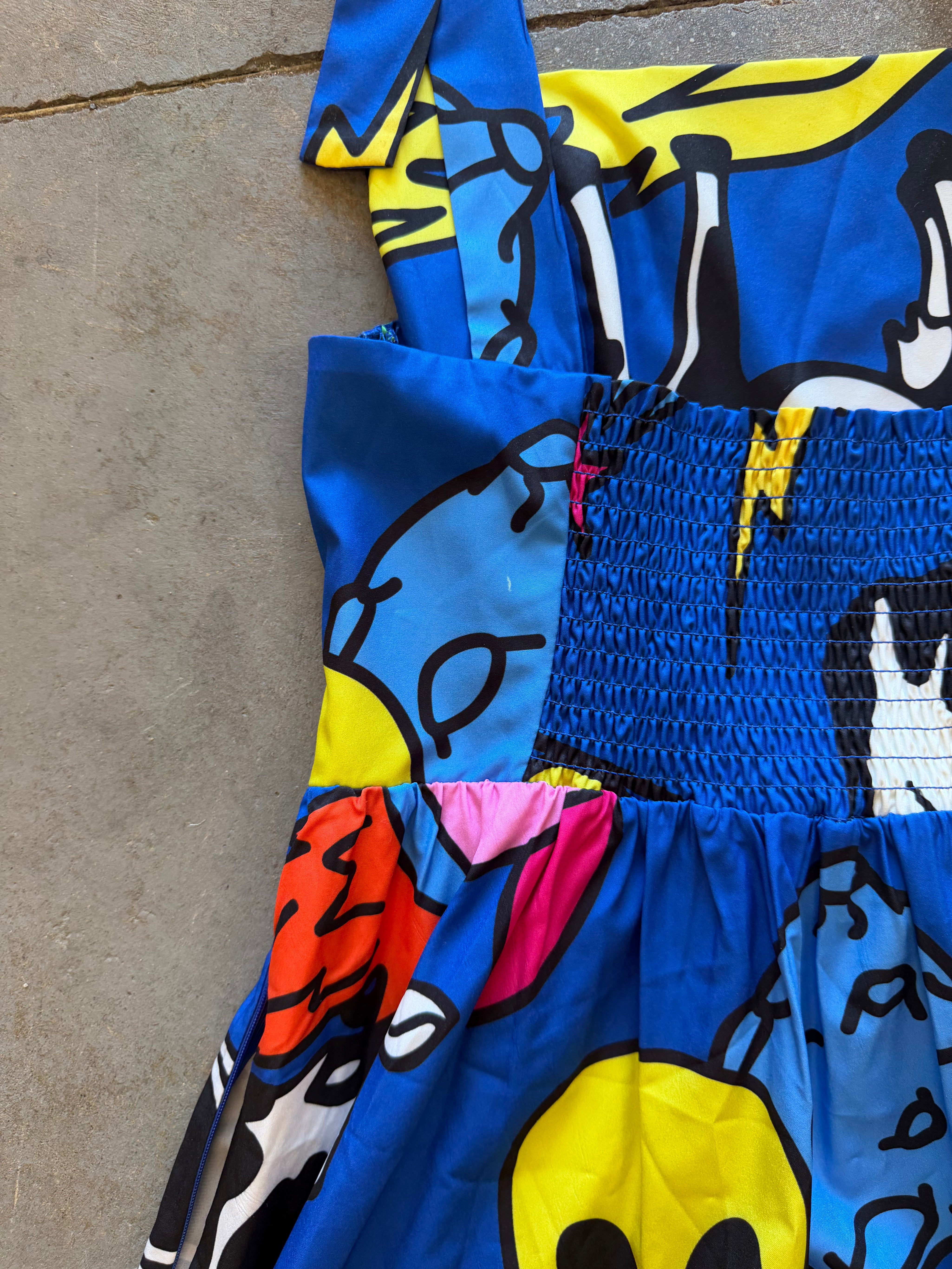 A close-up of a colorful printed garment laid flat on a concrete surface, featuring bold blue, yellow, black, pink, and orange abstract graphic patterns with a smocked waist section