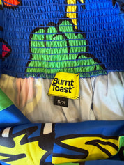 Close-up of a Burnt Toast garment with a bright blue smocked top, a green cartoon character graphic, and a yellow brand label reading 'Burnt Toast' with size 'S/M'