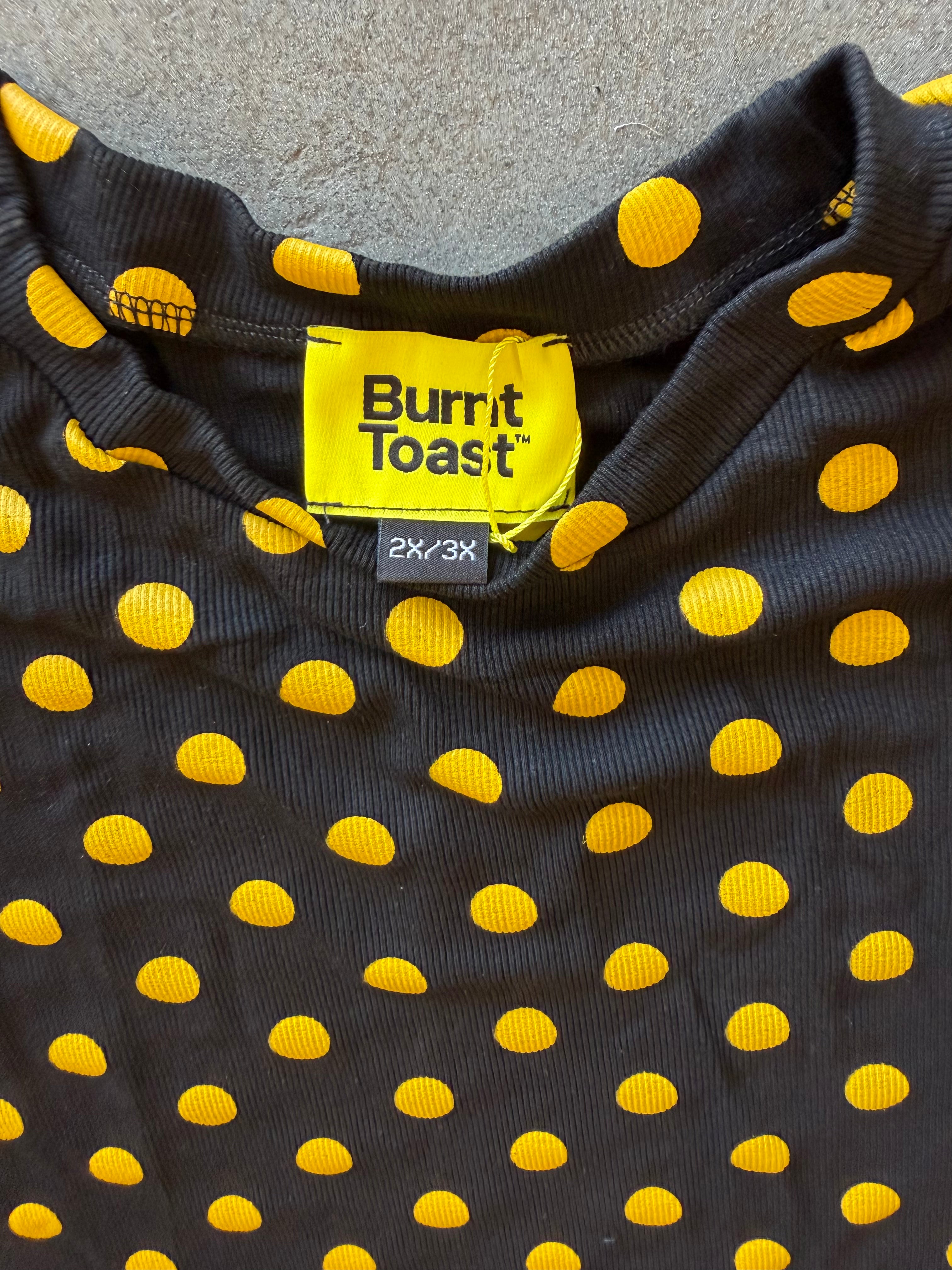 Close-up of a black ribbed knit garment with bright yellow polka dots, showing a yellow Burnit Toast neck label and a small 2X-3X size tag