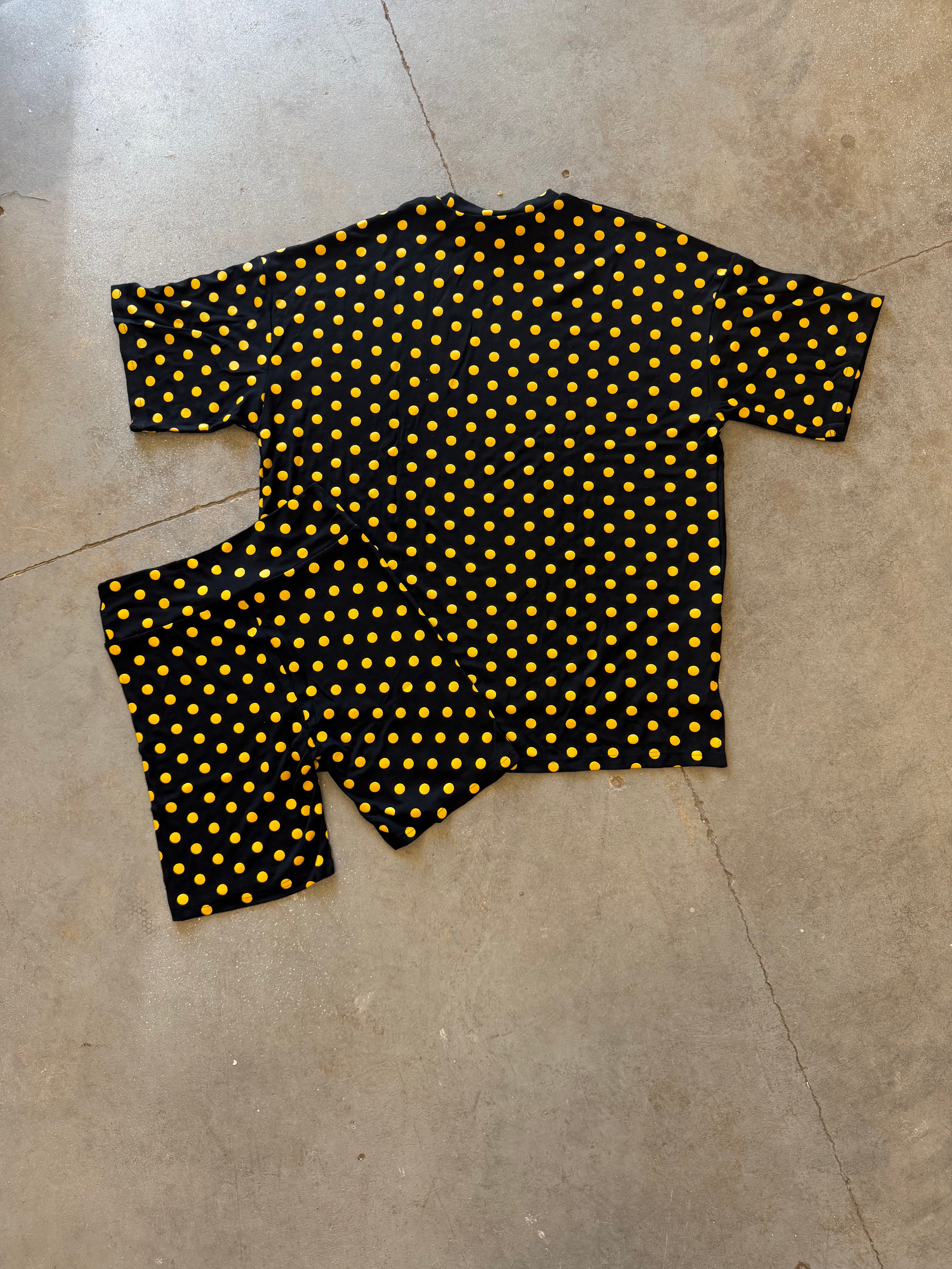 A two-piece matching set consisting of a short-sleeve top and shorts in black with a yellow polka dot pattern, laid flat on a concrete floor