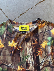 A close-up of a Burnt Toast crewneck top featuring a realistic autumn tree bark and maple leaf camouflage pattern, laid flat on a concrete surface