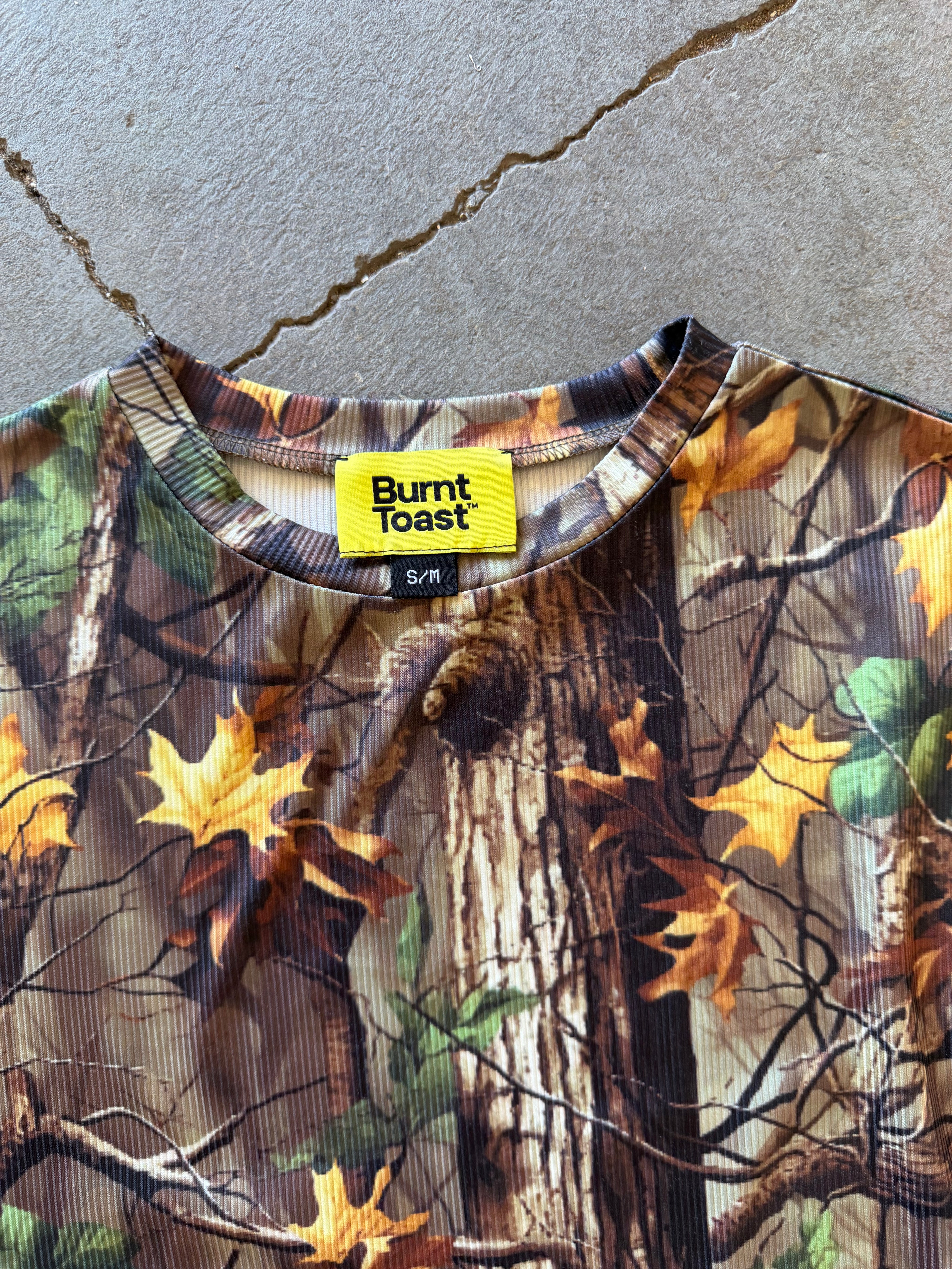 A close-up of a Burnt Toast crewneck top featuring a realistic autumn tree bark and maple leaf camouflage pattern, laid flat on a concrete surface