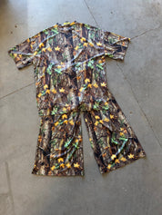 A short-sleeve camouflage ribbed set with matching shorts, laid flat on a gray concrete floor. The camouflage pattern features brown tree trunks, green leaves, and small yellow flowers