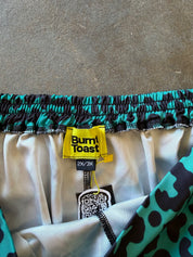 Close-up of a pair of shorts showing the elastic waistband with a black and teal pattern, a yellow Burn! Toast brand label, and a white satin inner lining