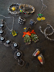 A flat-lay collection of gothic and Halloween-themed jewelry on a dark textured background, featuring silver-toned pieces with black, white, green, orange, yellow, and red details.