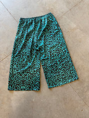 A pair of cropped pants with a vibrant turquoise and black leopard print pattern, laid flat on a beige concrete floor