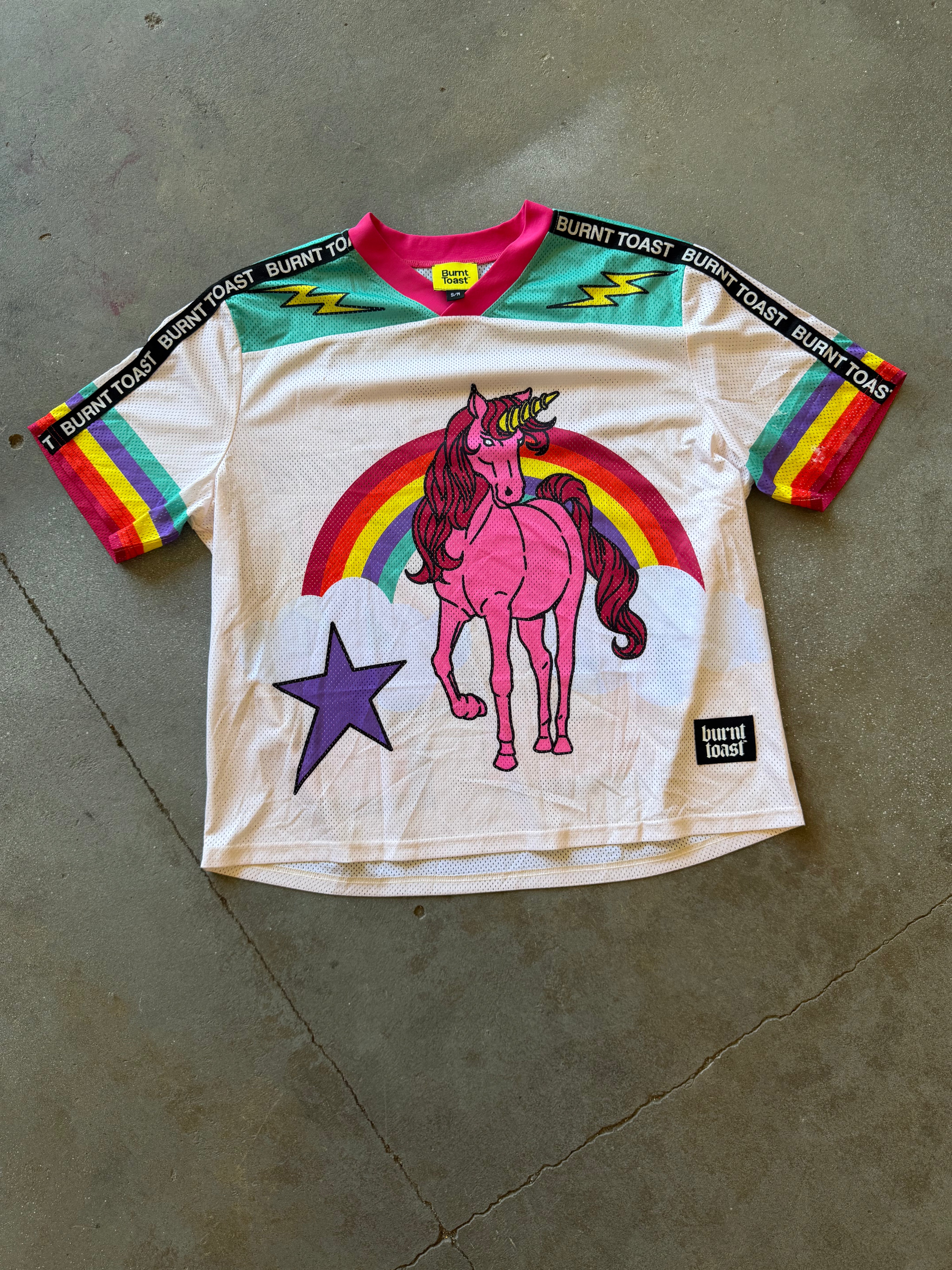 A white vintage-style Burnt Toast jersey laid flat on a concrete floor, featuring a pink unicorn under a rainbow graphic, a purple star, and rainbow-striped sleeves with 'BURNT TOAST' text across the shoulders.