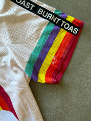 Close-up of a white mesh athletic jersey laid on a gray surface, featuring a black waistband with the words 'BURNT TOAST' in white and a vertical rainbow stripe pattern running down the side