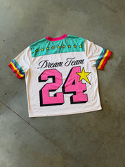A vintage-style white short-sleeve t-shirt with colorful graphics, laid flat on a gray concrete floor. The back of the shirt features 'Dream Team' in black script, a large pink number '24' with black and yellow outlines, a yellow star, and a teal chest band decorated with yellow stars