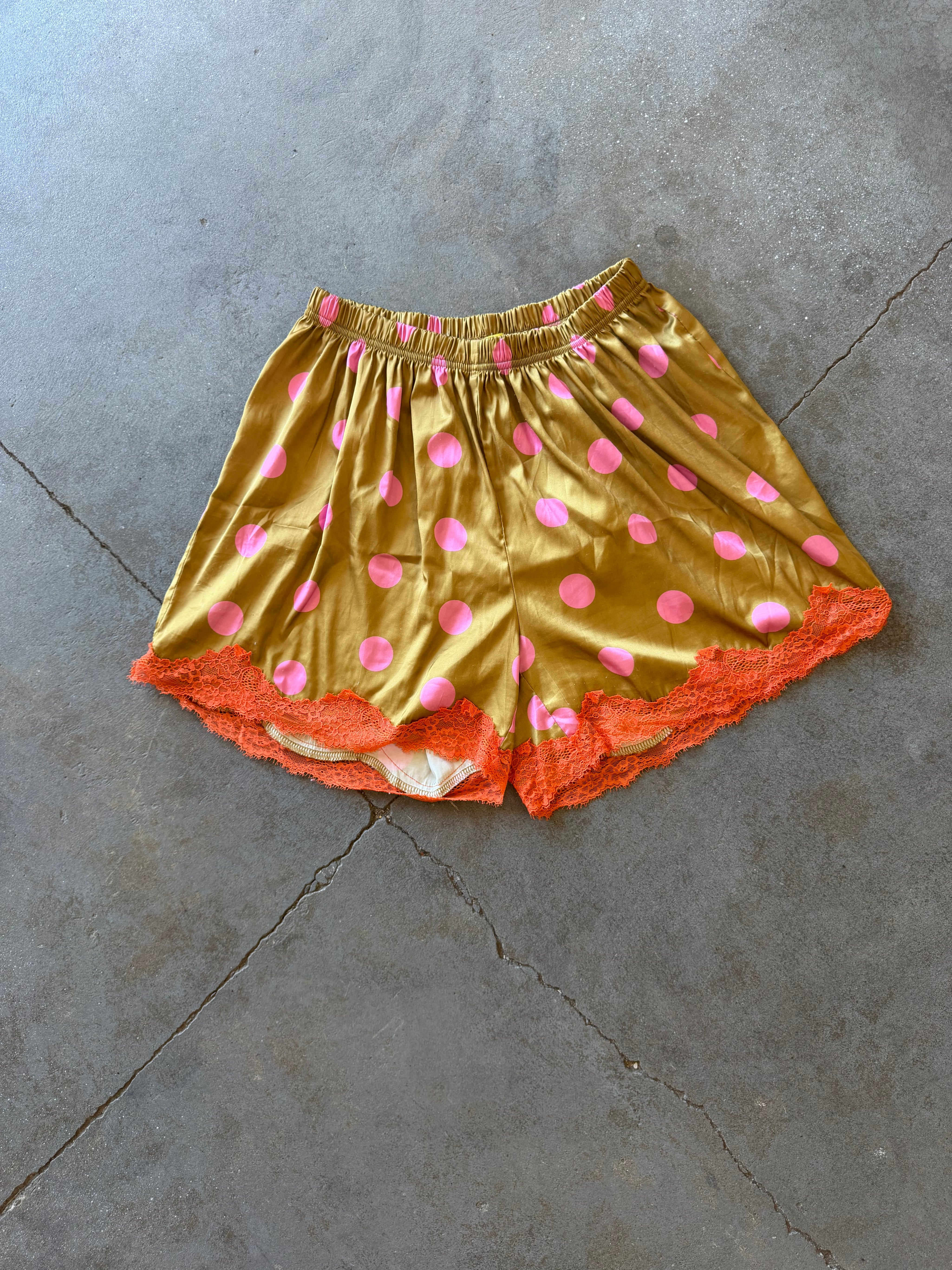 Gold satin shorts with pink polka dots and an orange lace hem, laid flat on a gray concrete floor