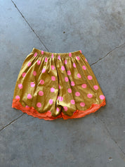 A pair of gold satin shorts with pink polka dots and orange lace trim, laid flat on a gray concrete floor