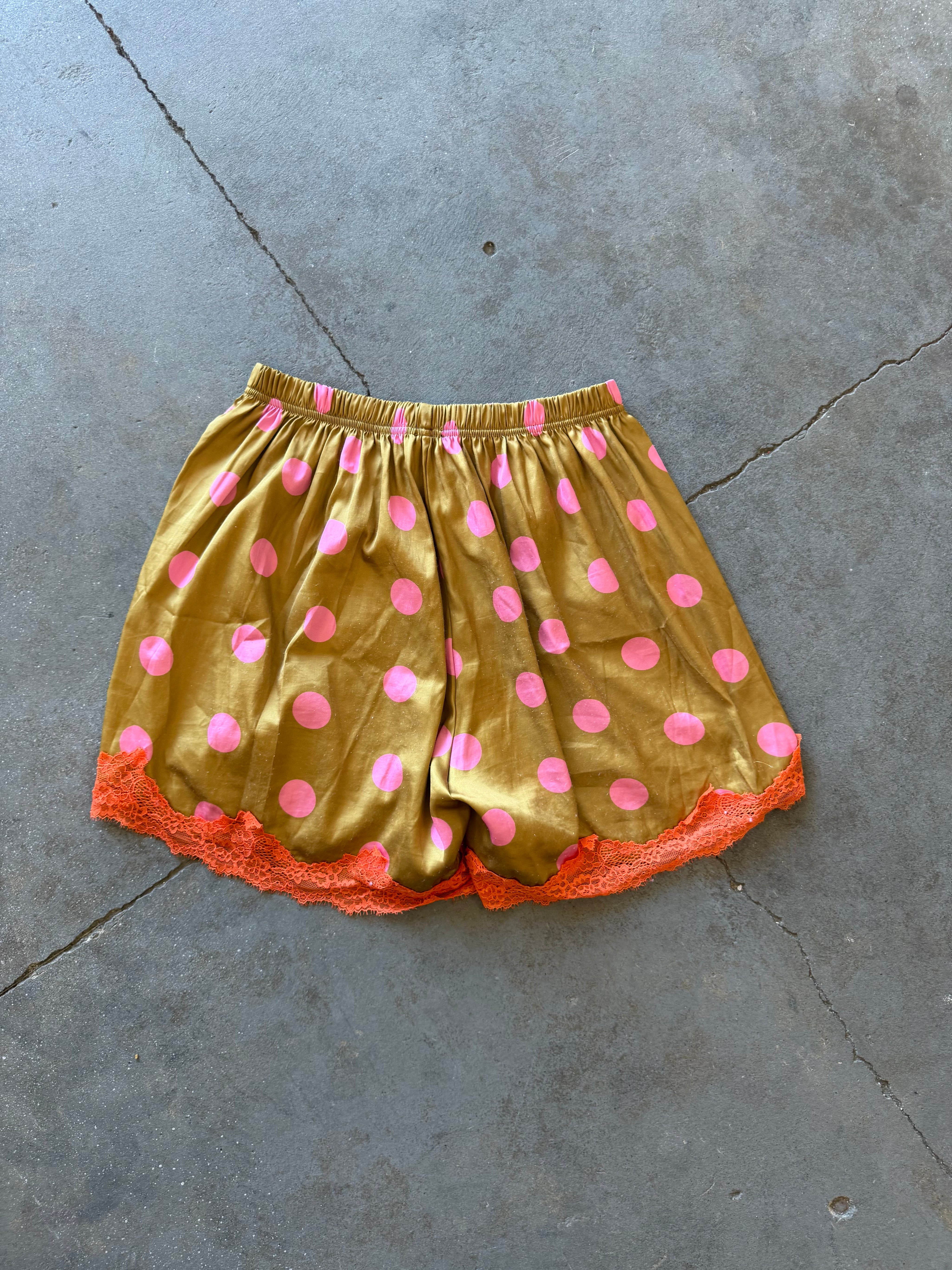 A pair of gold satin shorts with pink polka dots and orange lace trim, laid flat on a gray concrete floor