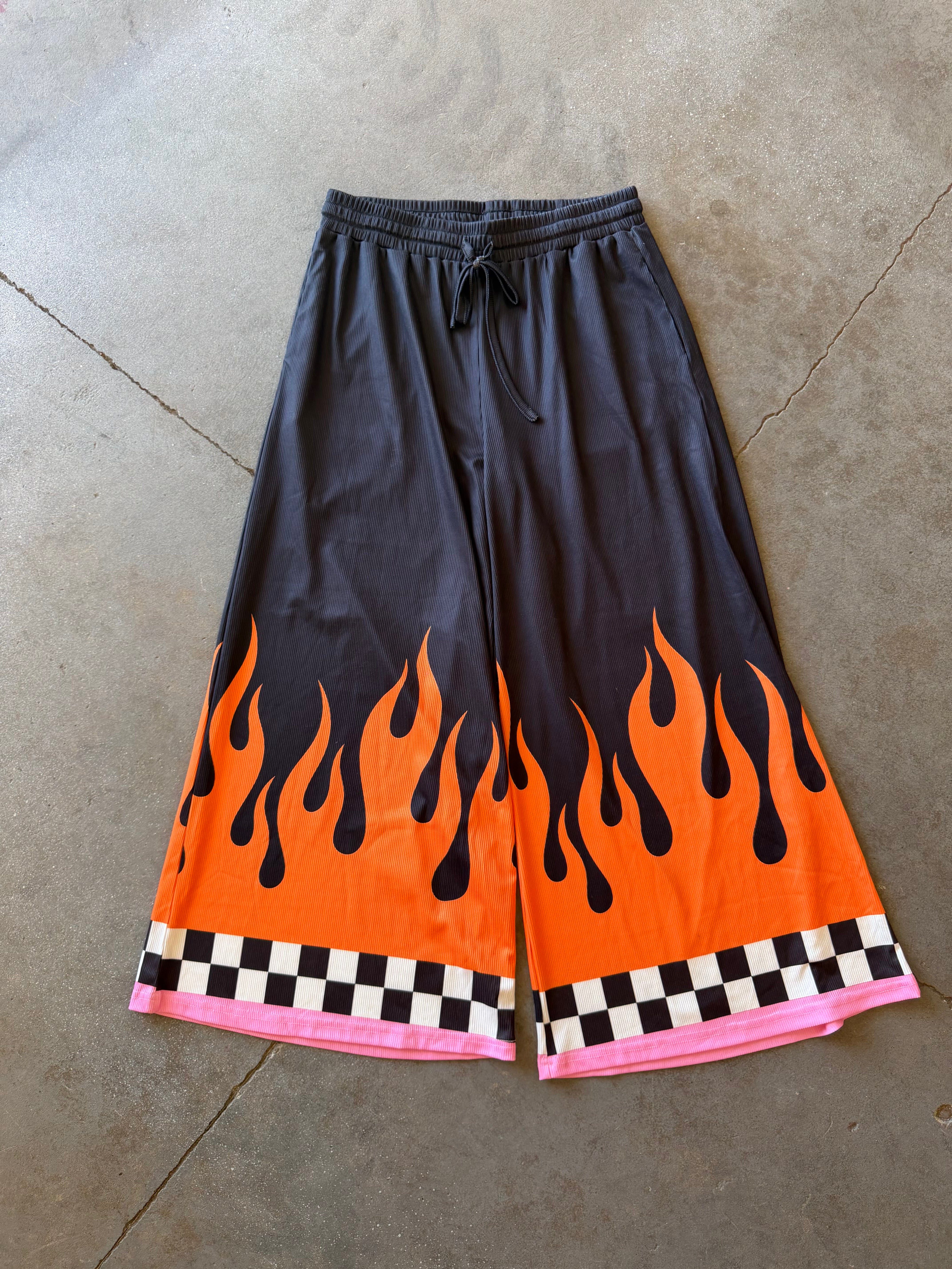 Black wide-leg shorts with a vibrant orange and black flame print near the hem, finished with a pink and black checkered border, laid flat on a concrete floor