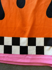 Close-up of a ribbed fabric garment showing a bright orange upper section with black teardrop-like shapes, a black-and-white checkerboard band, and a pink lower trim