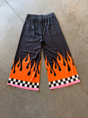 Black wide-leg athletic shorts with a vibrant orange and black flame print near the hem, finished with a pink and black checkered border, laid flat on a concrete floor