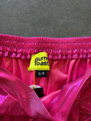 Close-up of a hot pink satin garment with a yellow Burnt Toast brand tag and black S/M size label, laid against a dark gray textured background